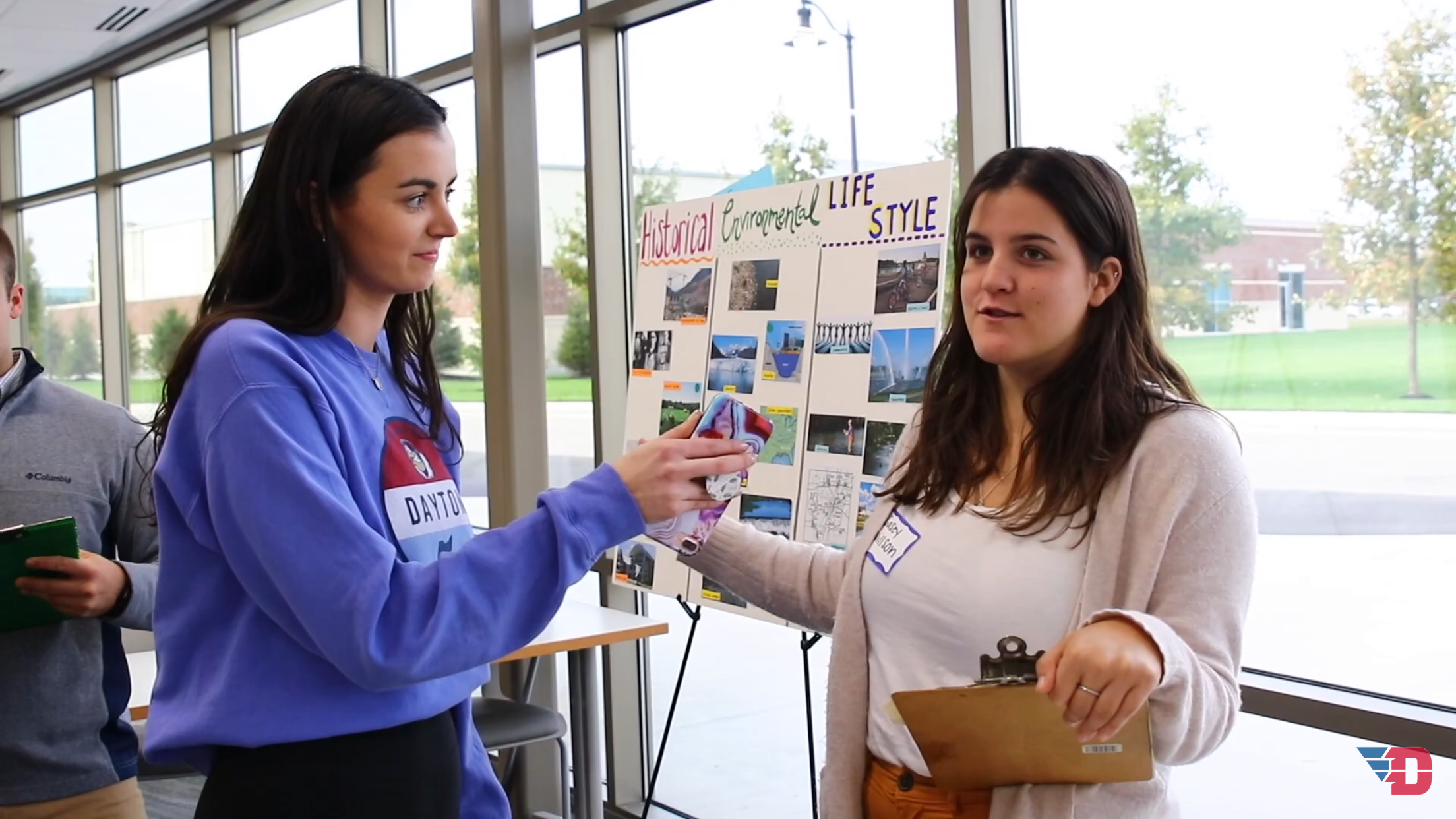 Image of a student interviewing another student with a presentation board in the background.