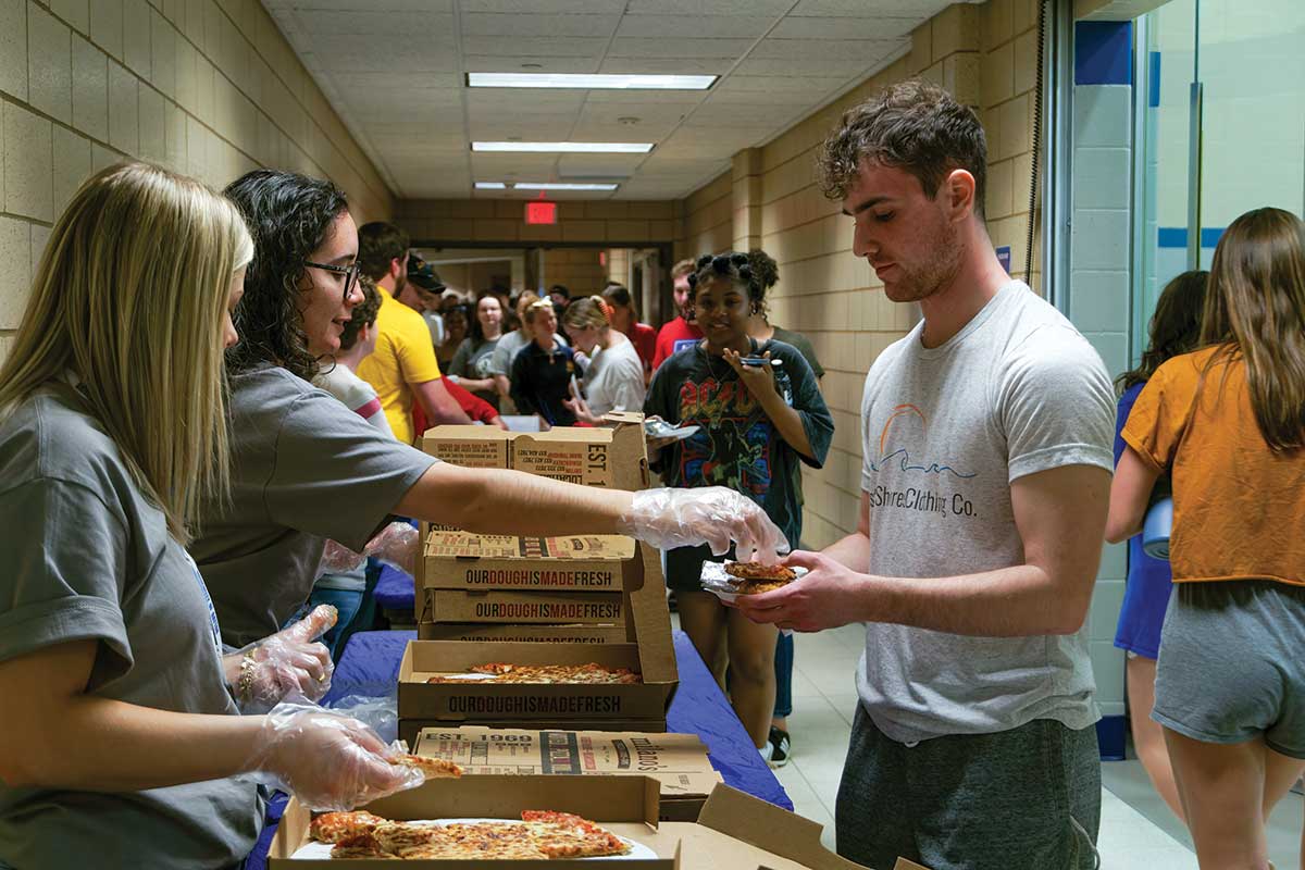 A long line os students waiting in front of tables to be served pizza.