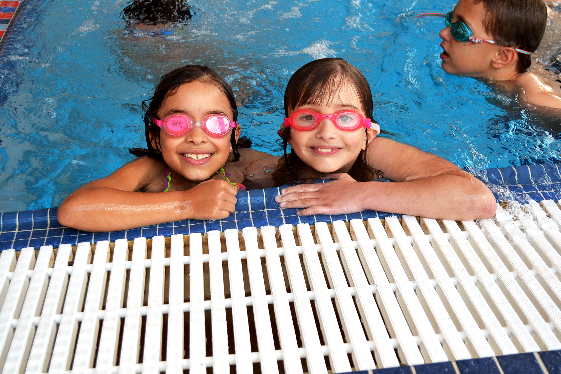 ${ Two young children wearing swim goggles and smiling while holding onto the side of the pool }