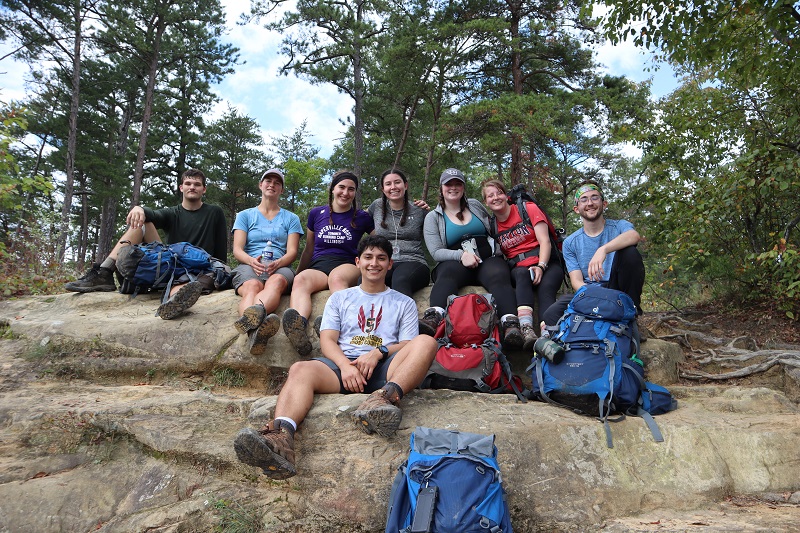 ${ Eight students on a backpacking trip smiling while sitting on a large rock. }